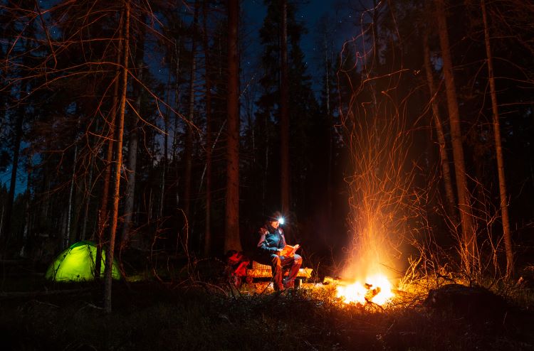 Woman reading a book with the light of a headlamp in the woods by a campfire